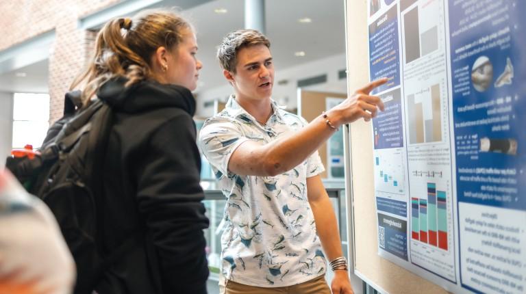 A student points to his research poster and discusses his findings with another student